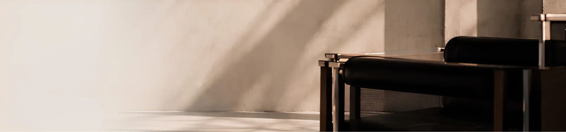 A dark armchair standing on a sunlit floor against a warm wall crossed by long diagonal shadows